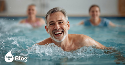 Homme d'âge moyen souriant lors d'un cours d'aquagym dans une piscine couverte, des éclaboussures d'eau autour de lui, deux autres participantes pratiquant des sports aquatiques en arrière-plan. L'image véhicule la joie, le mouvement et l'activité physique à un âge avancé.